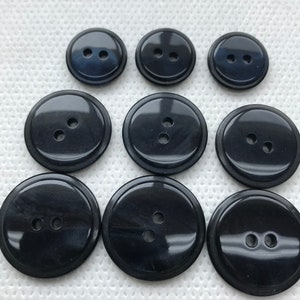 May include: Nine black buttons of varying sizes with two holes each. The buttons have a glossy finish and a slightly raised edge. The buttons are arranged on a white, textured surface.