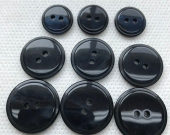 May include: Nine black buttons of varying sizes with two holes each. The buttons have a glossy finish and a slightly raised edge. The buttons are arranged on a white, textured surface.