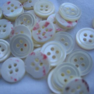 May include: A collection of off-white buttons with four holes each. Some buttons have a subtle pink and red speckled pattern. The buttons are round and have a slightly iridescent sheen, suggesting a mother-of-pearl or similar material.