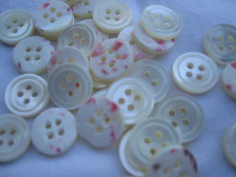 May include: A collection of off-white buttons with four holes each. Some buttons have a subtle pink and red speckled pattern. The buttons are round and have a slightly iridescent sheen, suggesting a mother-of-pearl or similar material.