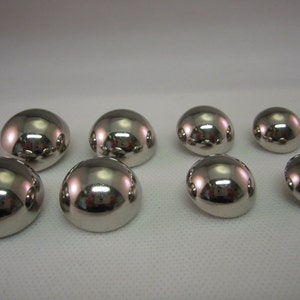 May include: Eight shiny, silver-coloured dome-shaped buttons. The buttons have a smooth, reflective surface, and vary slightly in size. They are arranged on a white surface, reflecting the surrounding environment.