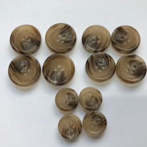 May include: A collection of ten round buttons in varying sizes. The buttons are a light brown color with a marbled, swirling pattern of darker brown. Each button has four holes for sewing.