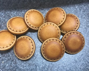 May include: A collection of eight round, light brown buttons with a stitched border. The buttons have a slightly textured surface and a darker brown stitched detail around the edge. They are arranged on a dark gray surface.