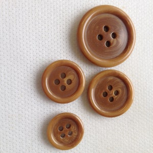May include: Four brown buttons of varying sizes with four holes each. The buttons have a glossy finish and a swirled pattern. The buttons are arranged on a white, textured surface.
