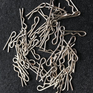 May include: A collection of silver cotter pins, also known as split pins, on a black background. These metal fasteners, with looped heads and split prongs, are used to secure mechanical parts.