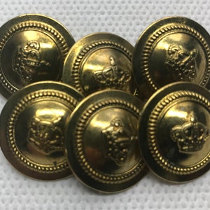 Pode incluir: Uma coleção de sete botões de latão antigos. Cada botão apresenta um design de coroa detalhado no centro, rodeado por uma borda de contas. Os botões têm uma aparência ligeiramente manchada e vintage.