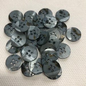 Puede incluir: Un conjunto de botones redondos de color gris oscuro, cada uno con cuatro agujeros. Los botones tienen un brillo sutil e iridiscente y están dispersos sobre una superficie blanca. Estos botones están hechos de un material duradero y son adecuados para varios proyectos de costura.