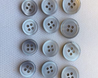 May include: A collection of twelve light gray buttons with four holes each. The buttons are arranged in three rows of four, with a slightly pearlescent finish. The buttons are round and have a subtle, iridescent sheen.