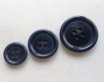 May include: Three navy blue buttons of varying sizes, each with four holes for sewing. The buttons have a marbled appearance and are arranged from smallest to largest, showcasing a range of sizes for different garment needs.