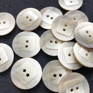 May include: A collection of round, off-white buttons with a square center. Each button has two holes for sewing. The buttons have a pearlescent sheen and a subtle, swirling pattern. These buttons are ideal for sewing projects.