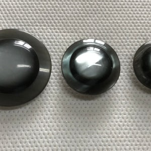 May include: Three dark gray, glossy buttons of varying sizes are arranged on a white, textured surface. The buttons have a slightly domed shape with a flat, circular rim. The largest button is approximately 1.5 inches in diameter.
