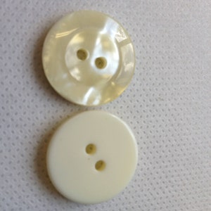 May include: Two round buttons. One is a pearlescent off-white with a raised center and two holes. The other is a solid off-white with two holes. Both buttons are for sewing and crafting.