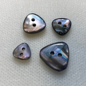 Pode incluir: Quatro botões triangulares iridescentes com dois furos cada. Os botões têm um brilho perolado com cores que variam do azul ao roxo e cinza. Os botões são de tamanhos variados.