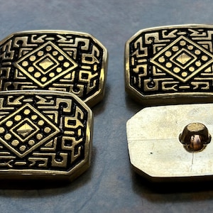 May include: Four antique brass buttons with an intricate geometric design. Each button is rectangular with a central diamond pattern surrounded by a series of lines and shapes. One button shows the button's back with a loop.