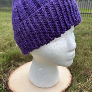 Puede incluir: Un gorro morado tejido con un diseño acanalado. El gorro se exhibe en una cabeza de maniquí blanca, apoyado sobre una rodaja de madera. El fondo presenta hierba verde y una valla.
