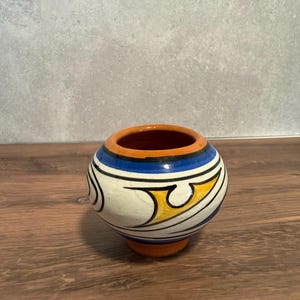 May include: A small, round ceramic pot with a wide opening, featuring a cream-colored body decorated with bold black, blue, and yellow abstract patterns. The rim and the base of the pot are finished in a solid, unglazed terracotta orange color.