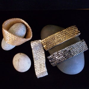 May include: Three woven bracelets in gold, silver, and black. The bracelets are made of small, square beads and are displayed on a gray rock.