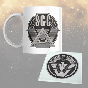 May include: White ceramic mug and glass coaster with a grey and black SGC logo design. The mug has a curved handle. The coaster has the SG logo with the number 1. The background has a soft, blurred effect.