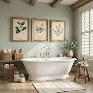 May include: A white freestanding bathtub with a silver faucet, positioned in a room with three framed botanical prints on the wall, woven wicker baskets containing rolled towels, a wooden stool, and glass jars on a stone tile floor.