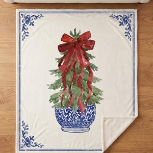 May include: A cream-coloured throw blanket featuring a watercolour illustration of a green pine tree in a blue and white patterned ceramic pot, topped with a large red bow. The blanket has a blue decorative border with floral corner accents.