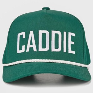 May include: A forest green baseball cap featuring the word CADDIE embroidered in bold white capital letters across the front panel. The cap includes a decorative white braided rope trim positioned horizontally along the base of the crown.