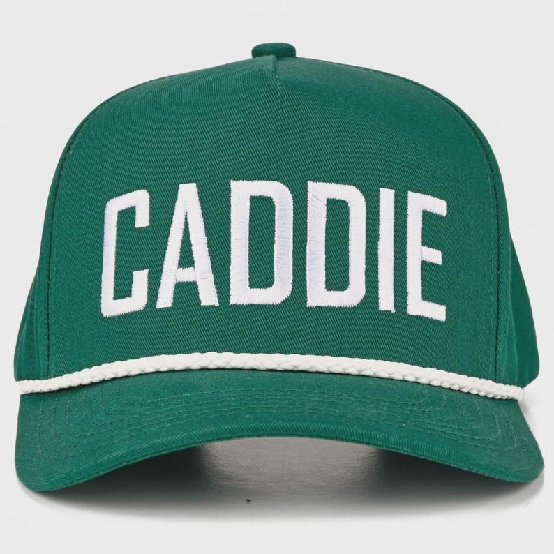 May include: A forest green baseball cap featuring the word CADDIE embroidered in bold white capital letters across the front panel. The cap includes a decorative white braided rope trim positioned horizontally along the base of the crown.