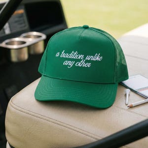 May include: A bright green trucker hat featuring a mesh back and a solid front panel. The text 'a tradition unlike any other' is embroidered in white cursive script across the center of the front panel. The hat has a curved brim and a button top.