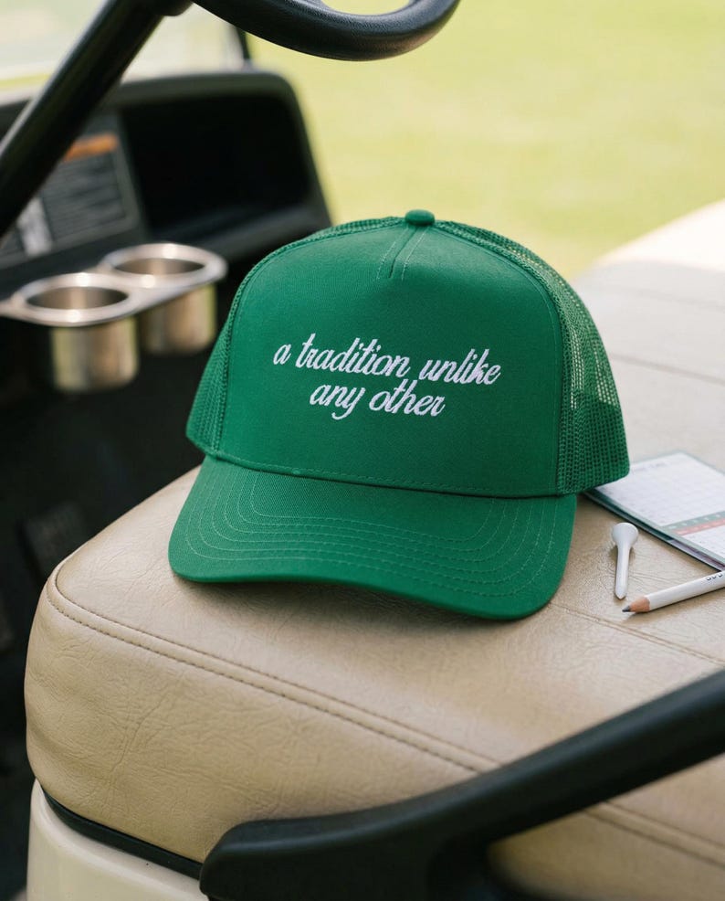 May include: A bright green trucker hat featuring a mesh back and a solid front panel. The text 'a tradition unlike any other' is embroidered in white cursive script across the center of the front panel. The hat has a curved brim and a button top.
