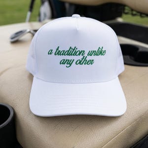May include: A white trucker hat featuring a structured front panel and a mesh back. The front of the hat displays the embroidered green text "a tradition unlike any other" in a cursive script font, centered above the solid white curved brim.