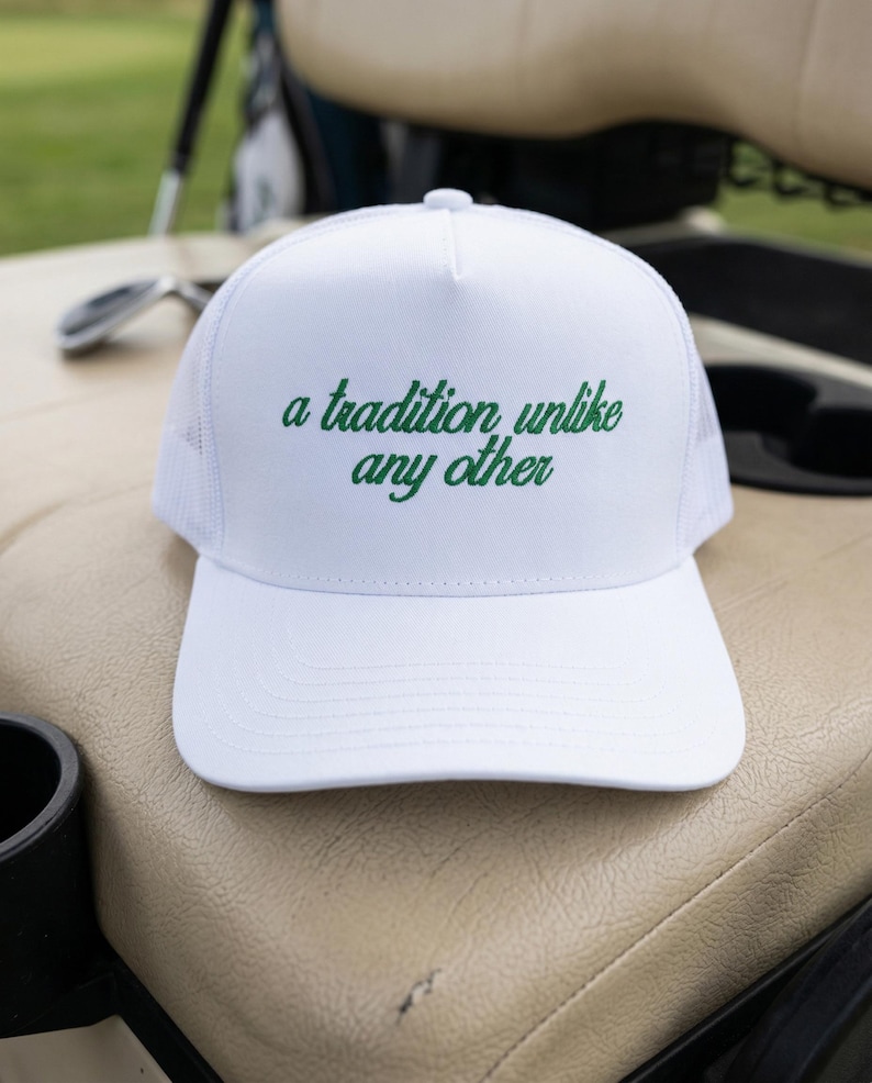 May include: A white trucker hat featuring a structured front panel and a mesh back. The front of the hat displays the embroidered green text "a tradition unlike any other" in a cursive script font, centered above the solid white curved brim.