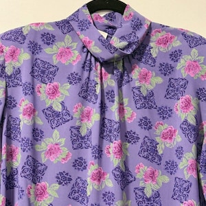 May include: A purple blouse with a high neckline, adorned with a repeating pattern of pink roses, green leaves, and dark purple geometric shapes. The blouse is hanging on a black hanger.