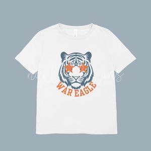 May include: White t-shirt featuring a tiger graphic with orange star-shaped sunglasses and the text "WAR EAGLE" in orange. The tiger is blue and white. The shirt is a classic crew neck.