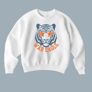 May include: White sweatshirt featuring a tiger graphic with orange star-shaped eyes and the text "WAR EAGLE" in orange. The tiger is blue and white. The sweatshirt has a crew neck and long sleeves.