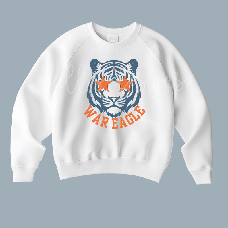 May include: White sweatshirt featuring a tiger graphic with orange star-shaped eyes and the text "WAR EAGLE" in orange. The tiger is blue and white. The sweatshirt has a crew neck and long sleeves.