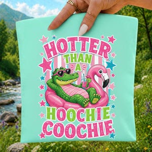 Peut inclure: Sac fourre-tout turquoise avec un motif d'alligator vert portant des lunettes de soleil en forme de cœur, se relaxant sur un flotteur de flamant rose. Le texte dit "HOTTER THAN A HOOCHIE COOCHIE" en rose et vert.