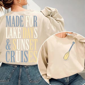 Peut inclure: Un sweat-shirt à col rond beige avec l'inscription MADE FOR LAKE DAYS & SUNSET CRUISES en bleu, blanc et jaune au dos. Le devant du sweat-shirt présente un petit motif représentant une pagaie jaune et bleue sur la poitrine gauche.