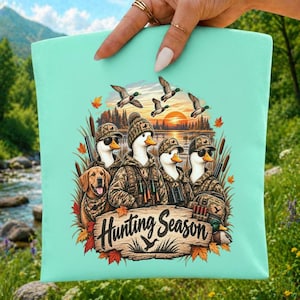 Peut inclure: Un sac fourre-tout vert menthe clair orné d'une illustration graphique de quatre canards portant des vêtements de chasse et des chapeaux de camouflage, accompagnés d'un golden retriever, avec le texte Hunting Season sur un panneau en bois.