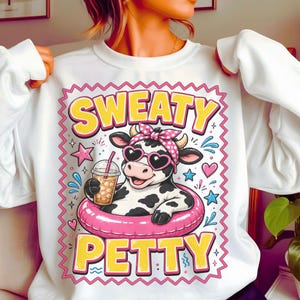 Peut inclure: Sweat-shirt blanc avec un dessin d'une vache dans une bouée rose, portant des lunettes de soleil en forme de cœur et un bandeau rose à pois. Le texte "SWEATY PETTY" est en jaune avec un bord rose. La vache tient une boisson avec une paille.