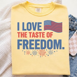 Peut inclure: Un t-shirt jaune pâle avec le texte bleu et rouge I LOVE THE TASTE OF FREEDOM. Le motif comprend un graphique stylisé du drapeau américain ainsi que de petites illustrations d'étoiles et de feux d'artifice rouges et bleus sur le devant.