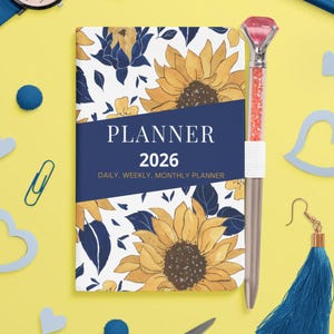 Peut inclure: Un agenda 2026 avec un motif de tournesol, un stylo avec un dessus en forme de diamant rose et une montre bleue sur fond jaune. L'agenda porte l'inscription "PLANNER 2026" et "DAILY, WEEKLY, MONTHLY PLANNER". Sont également inclus des ciseaux bleus, une boucle d'oreille à pompon bleu et un crayon bleu.