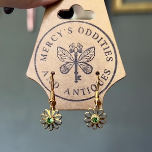 May include: A pair of gold-toned dangle earrings featuring a flower-shaped charm with a small green center stone. The earrings are attached to a tan cardboard display card printed with the text Mercy's Oddities and Antiques and a butterfly key.
