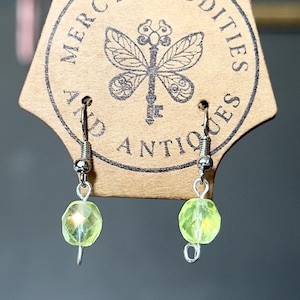 May include: A pair of dangle earrings featuring faceted, translucent lime green beads suspended from silver-toned metal hooks. The earrings are attached to a tan cardboard display card printed with the text Mercy's Oddities and Antiques.