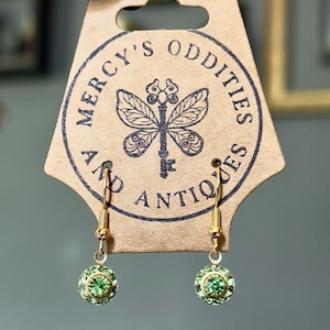 May include: A pair of gold-toned drop earrings featuring circular green crystal centers surrounded by small green rhinestones. The earrings are attached to a tan cardboard display card printed with the text Mercy's Oddities and Antiques.