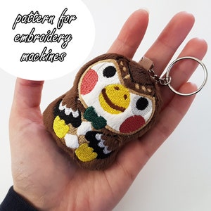 Puede incluir: Llavero de peluche marrón en forma de búho con detalles bordados, incluyendo una cara sonriente amarilla, una pajarita verde y detalles en blanco y negro. El llavero está unido a un anillo de metal plateado.