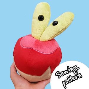 Puede incluir: Un juguete de peluche con forma de manzana roja con dos apéndices amarillos, peludos y con forma de gusano que sobresalen de la parte superior. El juguete tiene ojos negros y el texto "Sewing pattern" es visible en el fondo.