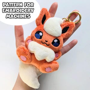 Pode incluir: Um porta-chaves de peluche laranja e creme com uma criatura estilizada de olhos azuis grandes, orelhas pontiagudas e uma cauda felpuda. O design inclui o texto PATTERN FOR EMBROIDERY MACHINES e está preso a um anel de metal dourado.