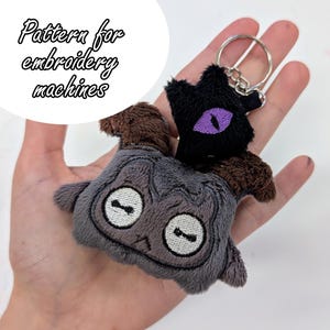 May include: A plush keychain featuring two cartoon creatures. One is gray with white eyes, and the other is black with a purple eye. The keychain has a silver ring. The text "Pattern for embroidery machines" is in the upper left corner.