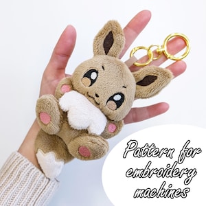 Op de afbeelding: Een bruine en witte Eevee pluche sleutelhanger met geborduurde gezichtskenmerken, roze pootkussentjes en een goudkleurige metalen ring. De afbeelding bevat de tekst Pattern for embroidery machines in een zwart lettertype op een witte achtergrond.