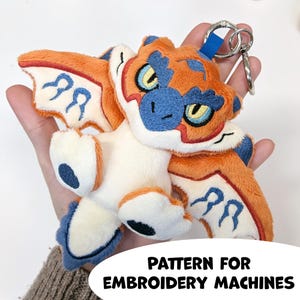 May include: A plush dragon keychain featuring orange, cream, and blue embroidered details with large yellow eyes and wings. The design includes a metal keyring attachment. The text at the bottom reads: PATTERN FOR EMBROIDERY MACHINES.