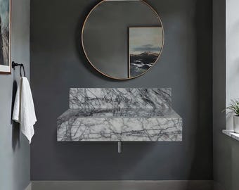 May include: A modern bathroom with a marble sink, a round gold-framed mirror, and a framed artwork. The sink has two faucets and a drain pipe. A white towel hangs on a black towel ring. The walls are painted a dark gray.